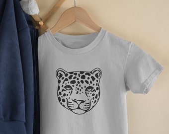May include: A light gray t-shirt featuring a black leopard face design. The leopard's face is centered on the shirt, with detailed spots and facial features. The shirt is hanging on a wooden hanger.