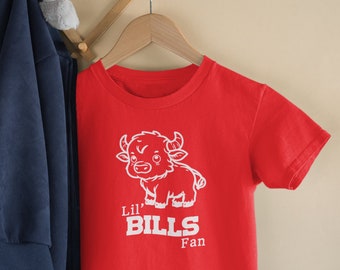 May include: A red t-shirt with a white cartoon buffalo graphic and the text "Lil' Bills Fan." The shirt is hanging on a wooden hanger. A blue jacket and a gray stuffed elephant are visible in the background.