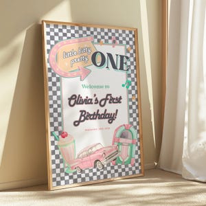 Puede incluir: Un letrero de cumpleaños enmarcado con un tema retro. El letrero presenta el texto "little bitty pretty ONE" y "Olivia's First Birthday!" junto con ilustraciones de un batido, un coche rosa y una gramola. El fondo tiene un patrón de cuadros blancos y negros.