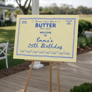 May include: A light yellow sign with blue text and a decorative border. The sign reads "Churing a year older & BUTTER than ever! Welcome to Emma's 25th Birthday." The sign is propped up on a wooden easel.