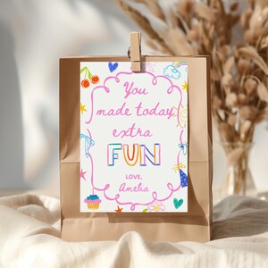 May include: A brown paper gift bag with a white card attached by a wooden clip. The card features colourful handwritten text saying "You made today extra FUN", with doodles of a rainbow, hearts, and a cupcake. The card is signed "Love, Amelia."