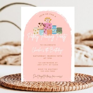 May include: A pink invitation for a pottery painting party. The invitation features a watercolor design with pottery and paintbrushes. Text includes "Pottery Painting Party" and event details. The invitation is on a woven placemat.