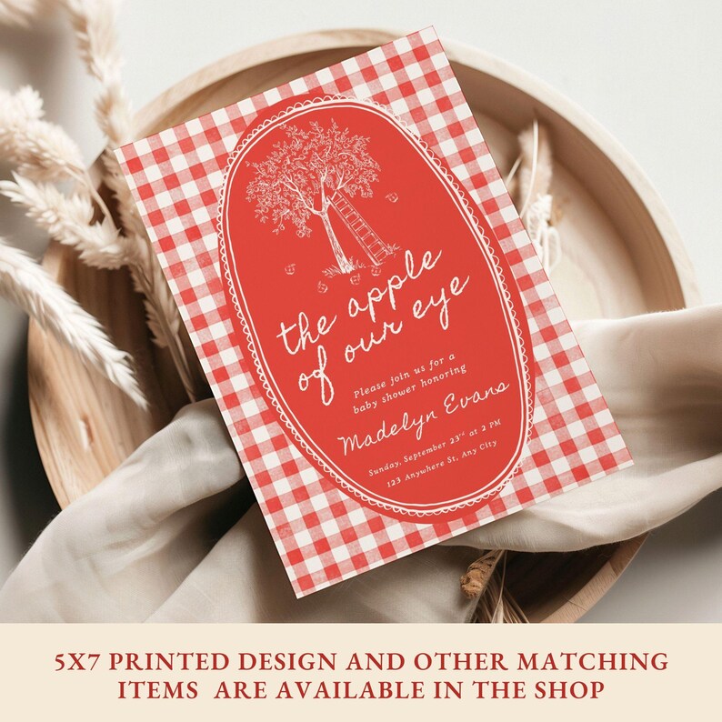 May include: A rectangular baby shower invitation card featuring a red and white gingham pattern. The center has a red oval frame containing a white line drawing of an apple tree with a ladder and the text: the apple of our eye, Please join us for a baby shower honoring Madelyn Evans, Sunday, September 23rd at 2 PM, 123 Anywhere St, Any City.
