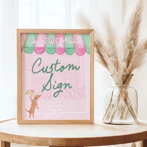 May include: A framed custom sign with a pink background, featuring the text "Cafe TWO Monde" and "Custom Sign" in green script. The sign includes a cartoon illustration of a dog holding a tray of pastries. The frame is made of light wood.