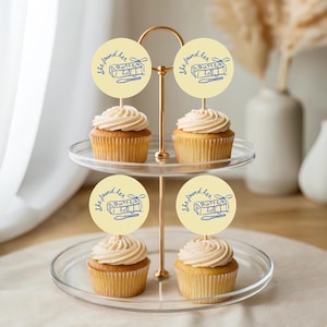 May include: A two-tiered glass dessert stand holds four cupcakes topped with swirled frosting. Each cupcake is adorned with a round, light yellow topper with the text "She found her Butter half" in blue. The stand has a gold-colored metal frame.