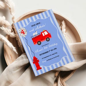 May include: A blue invitation card featuring a red fire engine, a Dalmatian, and a fire hydrant illustration. The text reads "STOP. DROP... and celebrate!" and "Oliver's 4th Birthday". The invitation is on a wooden tray with dried plants.