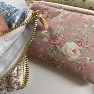 May include: Close-up of several floral print pouches with gold zippers. One pouch is open, revealing a white interior. The pouches feature pink, blue, and cream-colored fabrics with rose and other floral patterns. The zippers are a shiny gold color.