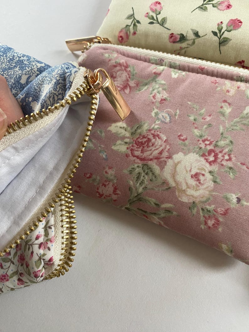 May include: Close-up of several floral print pouches with gold zippers. One pouch is open, revealing a white interior. The pouches feature pink, blue, and cream-colored fabrics with rose and other floral patterns. The zippers are a shiny gold color.