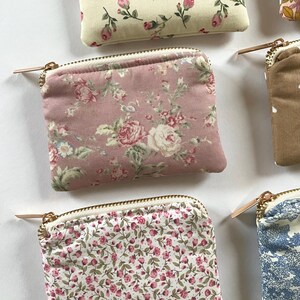 May include: Close-up of several small, square coin purses with floral patterns. The purses feature various designs, including pink roses, small pink flowers, and blue floral prints. Each purse has a gold zipper and a white interior.