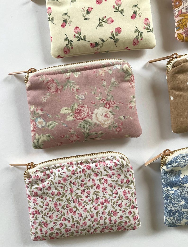 May include: Close-up of several small, square coin purses with floral patterns. The purses feature various designs, including pink roses, small pink flowers, and blue floral prints. Each purse has a gold zipper and a white interior.