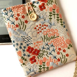 Könnte beinhalten: Ein beigefarbener Leinen-Buchbezug mit einem floralen Stickmuster. Die Stickerei zeigt rote, orange, rosa, blaue und grüne Blumen und Blätter. Ein Holz-Knopf mit vier Löchern ist an der Hülle befestigt.