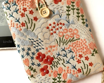 May include: A beige linen book cover with a floral embroidery design. The embroidery features red, orange, pink, blue, and green flowers and leaves. A wooden button with four holes is attached to the cover.