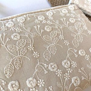 May include: Beige zippered pouch with intricate white floral embroidery. The design features detailed leaves, flowers, and vines, creating a delicate and elegant pattern. The pouch is made of a textured fabric.