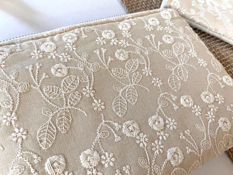May include: Beige zippered pouch with intricate white floral embroidery. The design features detailed leaves, flowers, and vines, creating a delicate and elegant pattern. The pouch is made of a textured fabric.
