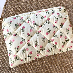 May include: Cream-colored quilted pouch with a floral pattern of pink roses and green leaves. The pouch has a diamond pattern and a gold zipper. The background is a woven, tan surface.