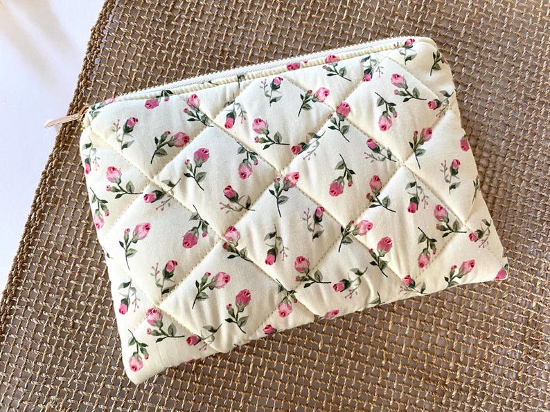May include: Cream-colored quilted pouch with a floral pattern of pink roses and green leaves. The pouch has a diamond pattern and a gold zipper. The background is a woven, tan surface.
