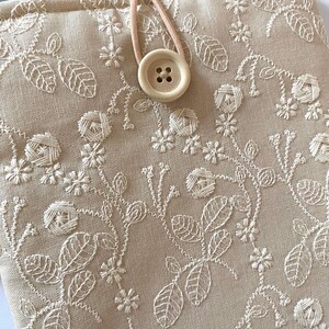Pu&ograve; includere: Una custodia per e-reader in lino beige con un ricamo floreale e una chiusura con bottone marrone.