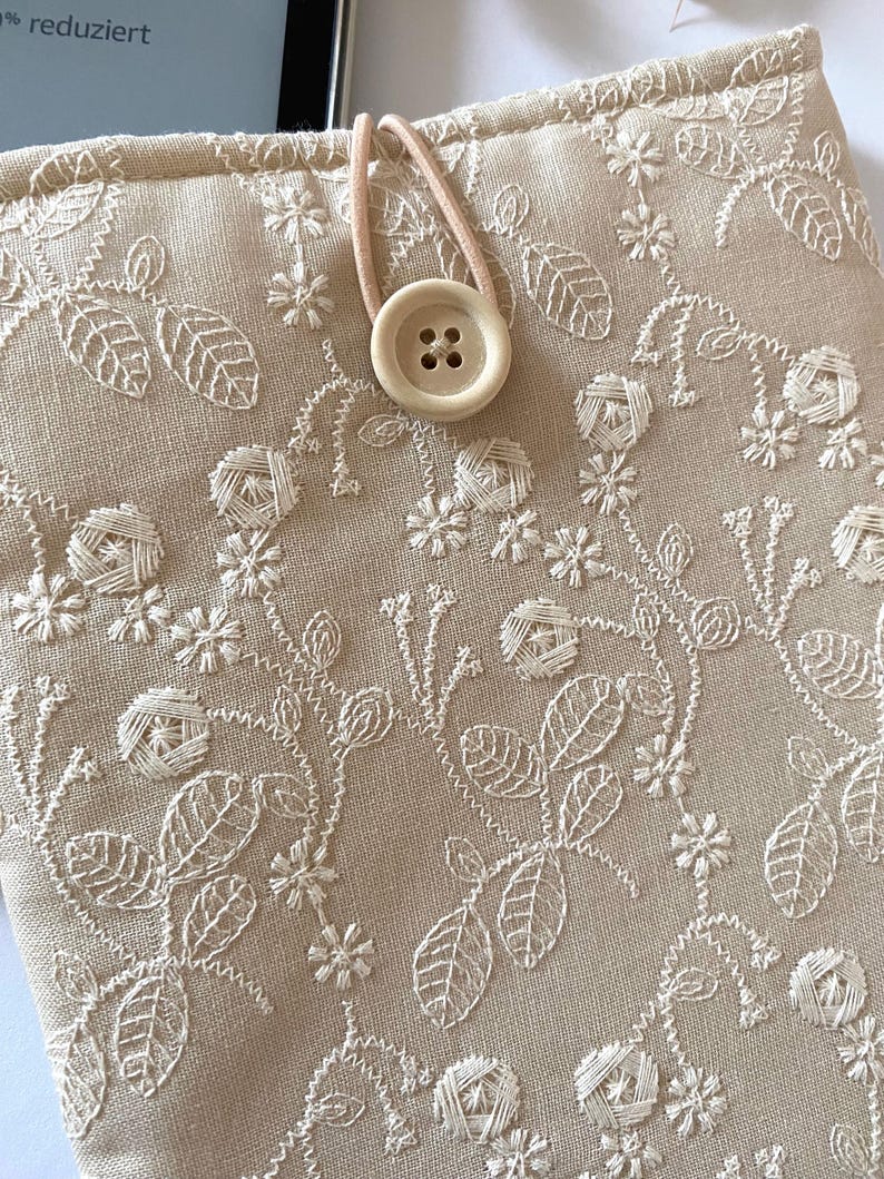 Pu&ograve; includere: Una custodia per e-reader in lino beige con un ricamo floreale e una chiusura con bottone marrone.