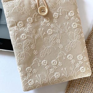 Pu&ograve; includere: Una custodia per e-reader in lino beige con un ricamo floreale bianco. La custodia ha una chiusura a bottone e un cinturino marrone.