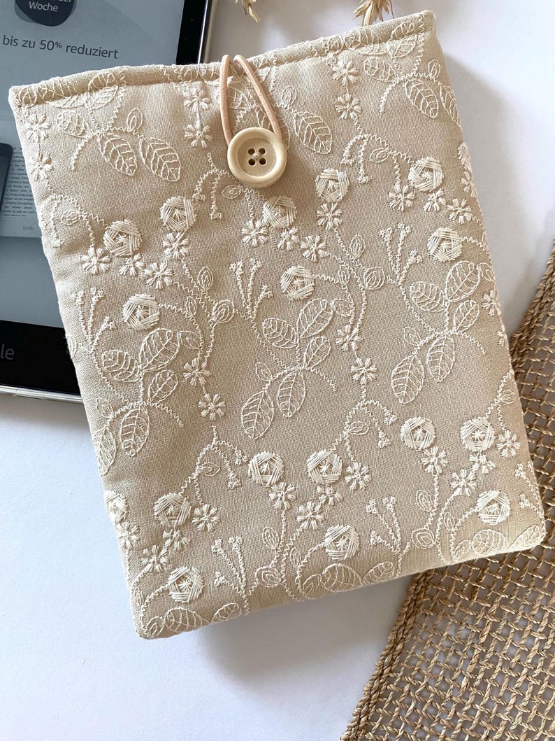 Pu&ograve; includere: Una custodia per e-reader in lino beige con un ricamo floreale bianco. La custodia ha una chiusura a bottone e un cinturino marrone.