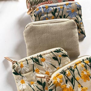 K&ouml;nnte beinhalten: Eine Sammlung kleiner Rei&szlig;verschlusstaschen. Einige sind aus beigem Leinen, w&auml;hrend andere mit gestickten Blumenmustern in verschiedenen Farben, darunter Rosa, Blau, Gelb und Wei&szlig;, versehen sind. Jede Tasche hat einen goldfarbenen Rei&szlig;verschluss und einen Zipper.