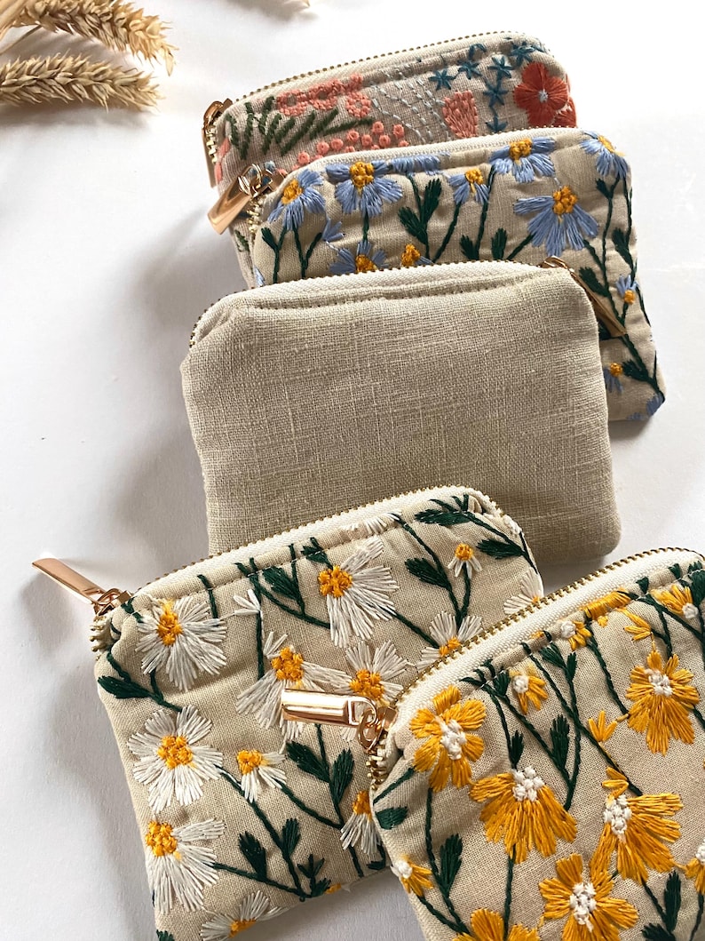 K&ouml;nnte beinhalten: Eine Sammlung kleiner Rei&szlig;verschlusstaschen. Einige sind aus beigem Leinen, w&auml;hrend andere mit gestickten Blumenmustern in verschiedenen Farben, darunter Rosa, Blau, Gelb und Wei&szlig;, versehen sind. Jede Tasche hat einen goldfarbenen Rei&szlig;verschluss und einen Zipper.