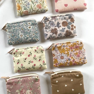 May include: A collection of small, rectangular coin purses with various floral and heart patterns. The purses feature a zipper closure with a gold-colored pull and are made of fabric. The color palette includes pink, yellow, blue, and brown.