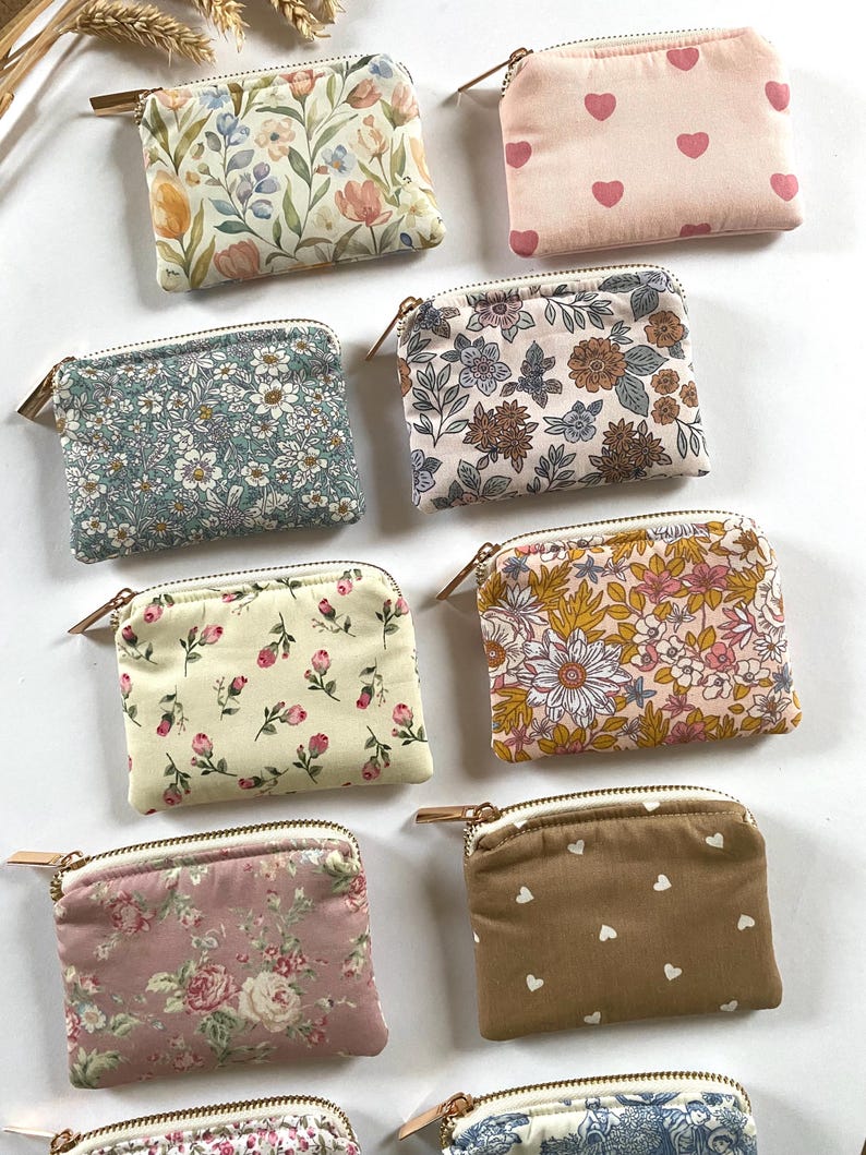 May include: A collection of small, rectangular coin purses with various floral and heart patterns. The purses feature a zipper closure with a gold-colored pull and are made of fabric. The color palette includes pink, yellow, blue, and brown.