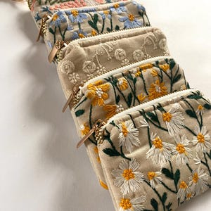 K&ouml;nnte beinhalten: Eine Reihe beigefarbener Stoffbeutel mit floralen Stickereien. Jeder Beutel zeigt ein anderes gesticktes Blumendesign in verschiedenen Farben, darunter Rosa, Blau, Gelb und Wei&szlig;. Goldfarbene Rei&szlig;verschl&uuml;sse.