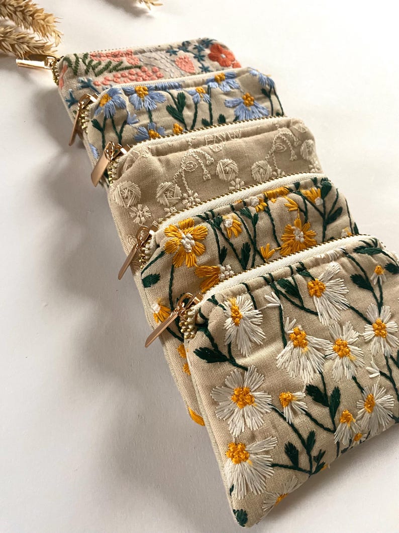K&ouml;nnte beinhalten: Eine Reihe beigefarbener Stoffbeutel mit floralen Stickereien. Jeder Beutel zeigt ein anderes gesticktes Blumendesign in verschiedenen Farben, darunter Rosa, Blau, Gelb und Wei&szlig;. Goldfarbene Rei&szlig;verschl&uuml;sse.
