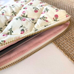 May include: Cream-colored quilted case with a floral pattern of pink roses and green leaves. The case has a gold zipper and a pink interior. The design is reminiscent of vintage floral patterns.
