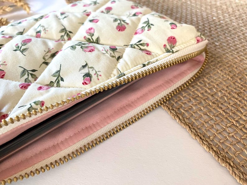 May include: Cream-colored quilted case with a floral pattern of pink roses and green leaves. The case has a gold zipper and a pink interior. The design is reminiscent of vintage floral patterns.