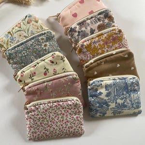 May include: A collection of small, rectangular coin purses with various floral and patterned designs. The purses feature gold-colored zippers and are arranged in a neat stack. Colors include pink, blue, white, and brown.