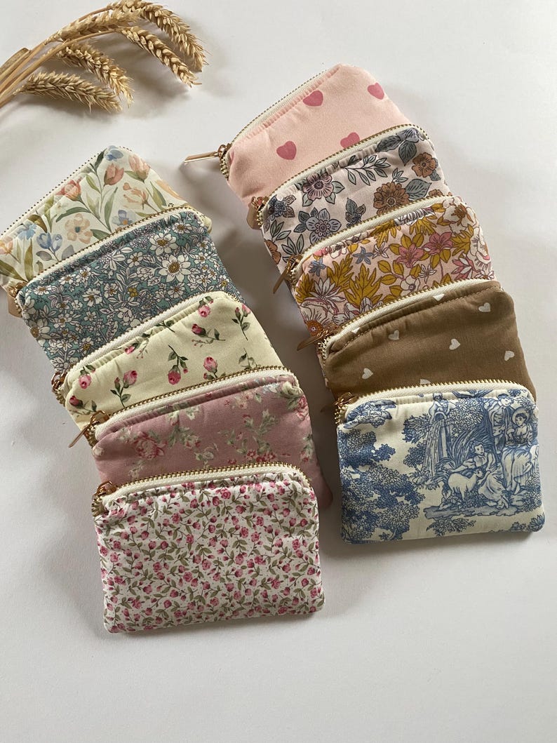 May include: A collection of small, rectangular coin purses with various floral and patterned designs. The purses feature gold-colored zippers and are arranged in a neat stack. Colors include pink, blue, white, and brown.