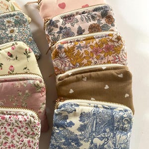 May include: A collection of small, zippered pouches in various floral and patterned fabrics. The pouches feature gold-colored zippers and a variety of colors, including pink, blue, and brown. Some pouches have heart patterns, while others display detailed floral designs.