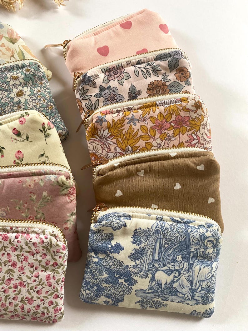 May include: A collection of small, zippered pouches in various floral and patterned fabrics. The pouches feature gold-colored zippers and a variety of colors, including pink, blue, and brown. Some pouches have heart patterns, while others display detailed floral designs.