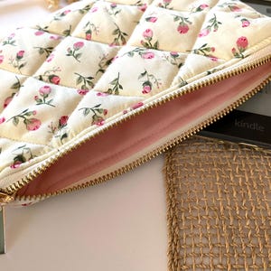 May include: Cream-colored quilted case with a floral pattern of pink roses and green leaves. The case has a gold zipper and a soft pink interior. The word "kindle" is visible on a black electronic device.