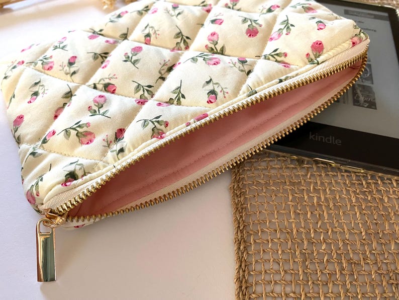 May include: Cream-colored quilted case with a floral pattern of pink roses and green leaves. The case has a gold zipper and a soft pink interior. The word "kindle" is visible on a black electronic device.