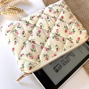 May include: Cream-colored quilted case with a floral pattern of pink roses and green leaves. The case has a gold zipper and is partially open, revealing an e-reader. The case is designed to protect an e-reader.