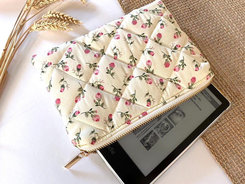 May include: Cream-colored quilted case with a floral pattern of pink roses and green leaves. The case has a gold zipper and is partially open, revealing an e-reader. The case is designed to protect an e-reader.