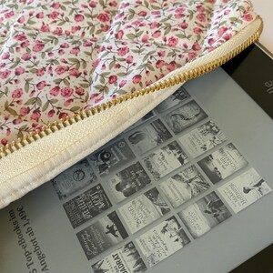 May include: A white quilted e-reader case with a floral print and a gold zipper. The case is open and a Kindle e-reader is inside. The Kindle screen shows a grid of book covers with titles like "The Empty Man" and "The Night Circus".