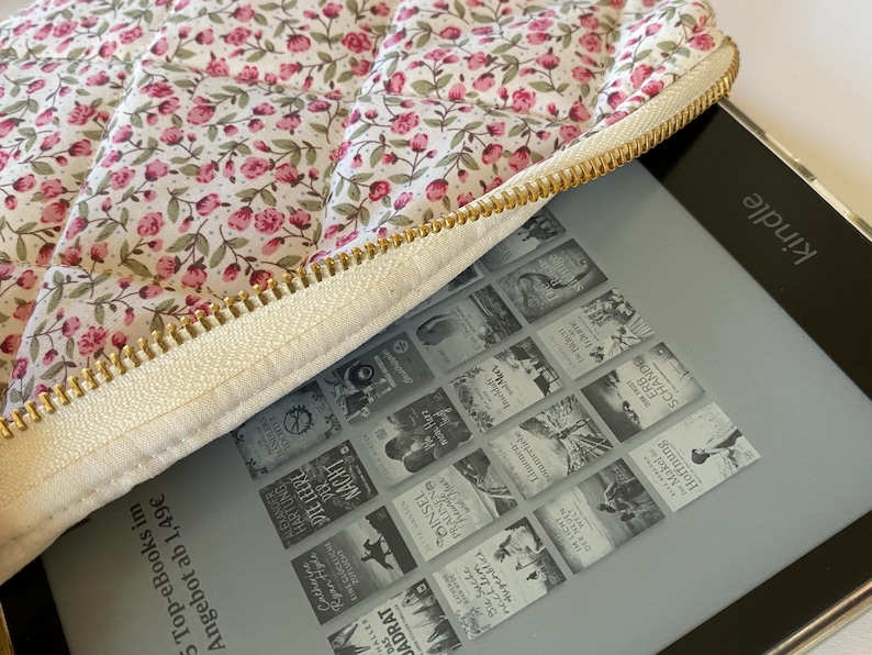 May include: A white quilted e-reader case with a floral print and a gold zipper. The case is open and a Kindle e-reader is inside. The Kindle screen shows a grid of book covers with titles like "The Empty Man" and "The Night Circus".