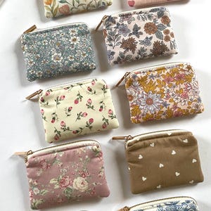 Könnte beinhalten: Eine Sammlung kleiner, quadratischer Stoffbeutel mit Reißverschlüssen. Die Beutel weisen verschiedene Blumen- und Musterdesigns in Rosa, Blau, Gelb und Braun auf. Einige haben Herzmuster. Jeder Beutel hat einen goldfarbenen Reißverschluss und einen kleinen Lederzug.