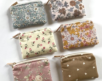 May include: A collection of small, square fabric pouches with zippers. The pouches feature various floral and patterned designs in shades of pink, blue, yellow, and brown. Some have heart patterns. Each pouch has a gold-colored zipper and a small leather pull.