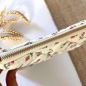 May include: Cream-colored quilted pouch with a floral pattern of pink roses and green leaves. The pouch has a gold zipper closure and a matching gold zipper pull. The overall theme is feminine and romantic.