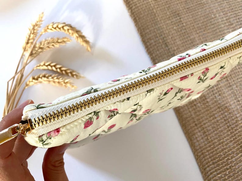 May include: Cream-colored quilted pouch with a floral pattern of pink roses and green leaves. The pouch has a gold zipper closure and a matching gold zipper pull. The overall theme is feminine and romantic.