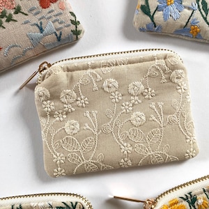 K&ouml;nnte beinhalten: Beige bestickte M&uuml;nzb&ouml;rsen mit floralen Designs. Die Geldb&ouml;rsen weisen aufwendige wei&szlig;e, gelbe, blaue und orangefarbene Blumenstickereien auf. Jede Geldb&ouml;rse hat einen goldenen Rei&szlig;verschluss und eine rechteckige Form.