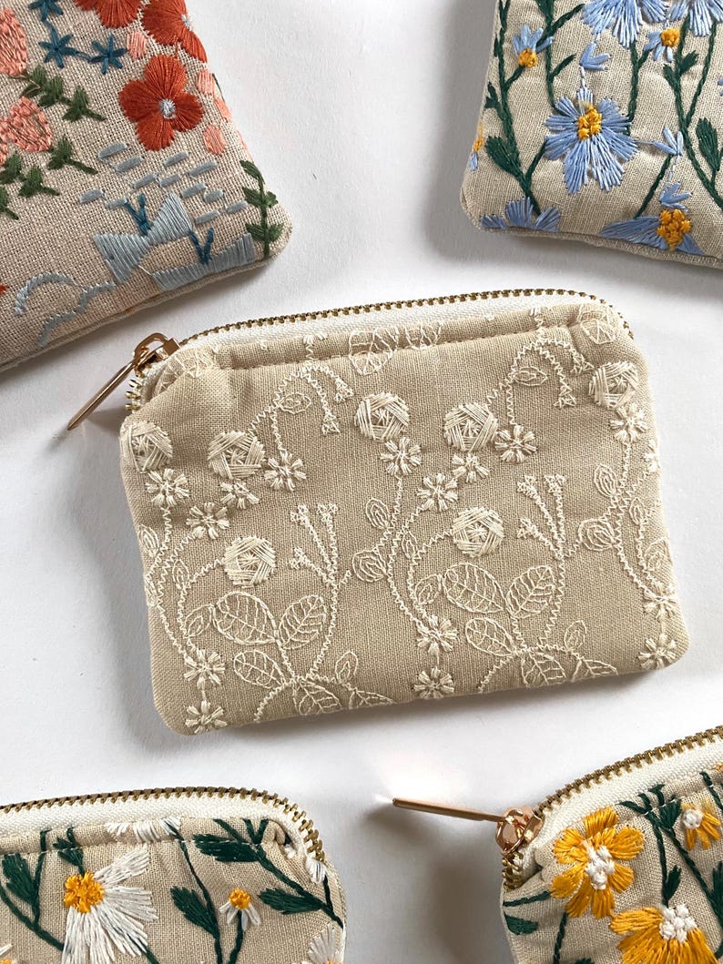 K&ouml;nnte beinhalten: Beige bestickte M&uuml;nzb&ouml;rsen mit floralen Designs. Die Geldb&ouml;rsen weisen aufwendige wei&szlig;e, gelbe, blaue und orangefarbene Blumenstickereien auf. Jede Geldb&ouml;rse hat einen goldenen Rei&szlig;verschluss und eine rechteckige Form.
