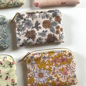May include: A collection of small, zippered pouches with floral patterns. The pouches feature various designs, including flowers, leaves, and hearts, in colors like pink, yellow, blue, and brown. Each pouch has a gold-colored zipper.