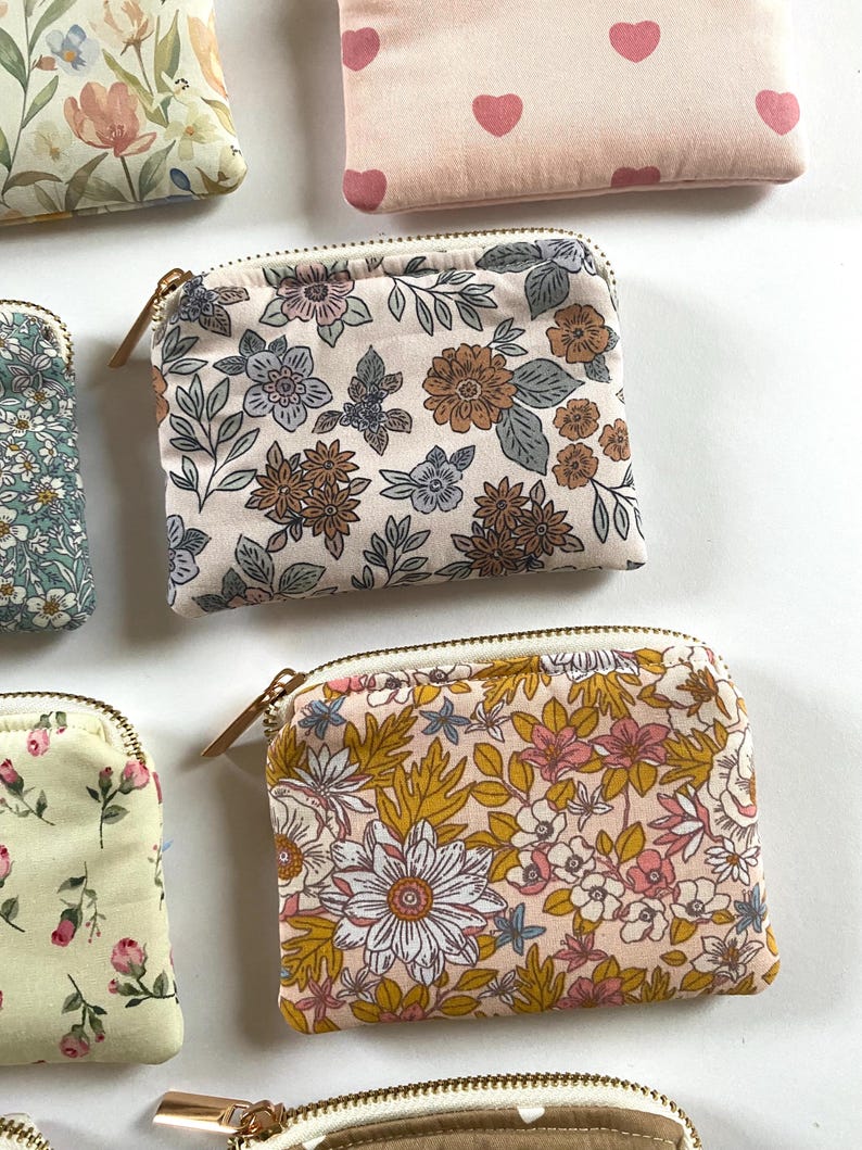 May include: A collection of small, zippered pouches with floral patterns. The pouches feature various designs, including flowers, leaves, and hearts, in colors like pink, yellow, blue, and brown. Each pouch has a gold-colored zipper.